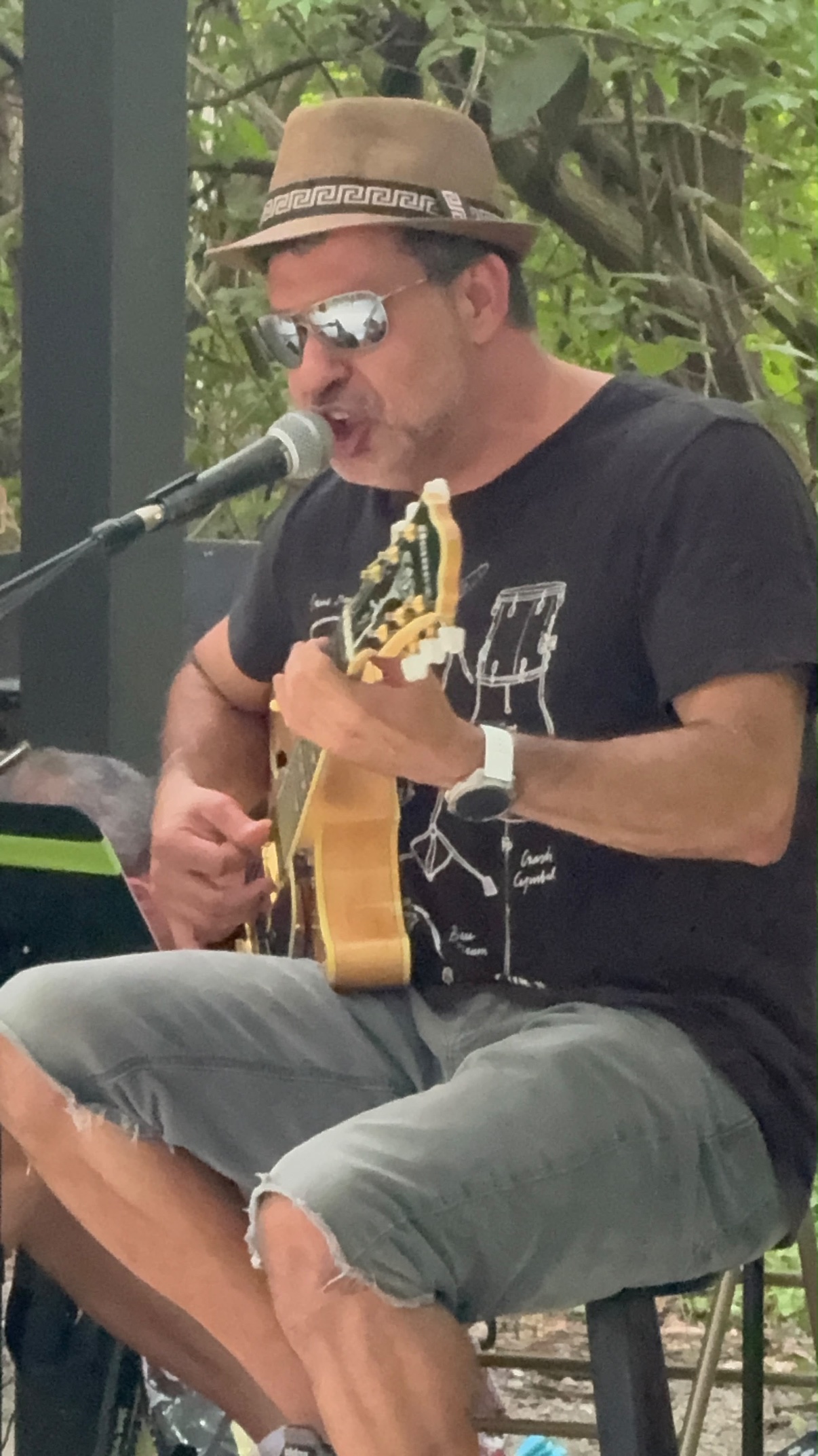 Doug Dantas singing into a microphone while playing guitar outdoors, wearing a hat and sunglasses